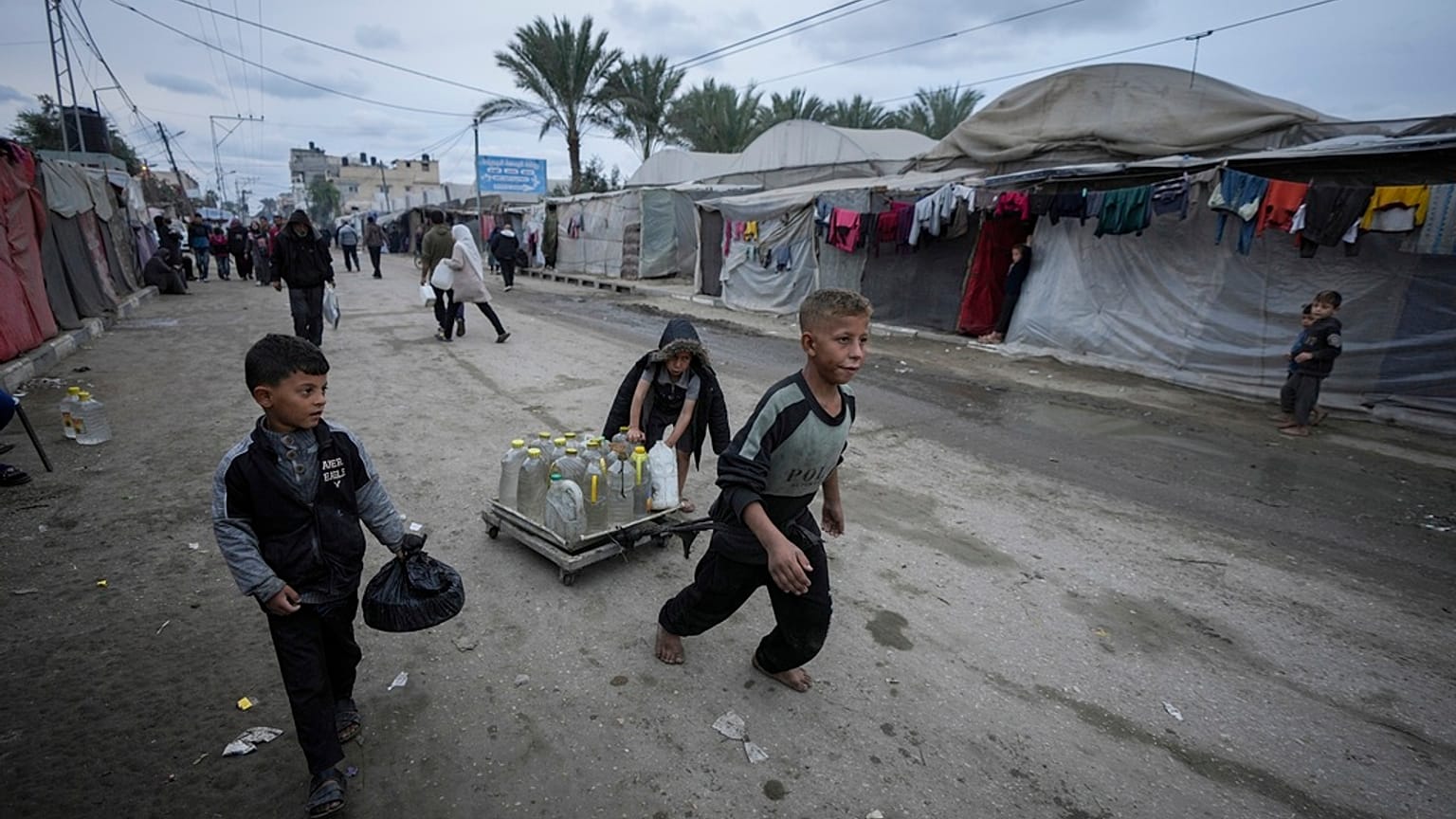 أطفال فلسطينيون يدفعون عربة تحمل صفائح المياه وزجاجات المياه البلاستيكية في مخيم للنازحين في دير البلح بقطاع غزة