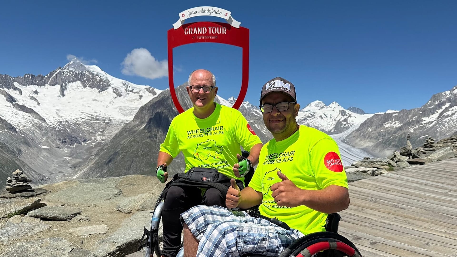 Two smiling men in wheelchairs wearing bright yellow shirts in the foreground, a red sign and snowy mountains in the background. 
