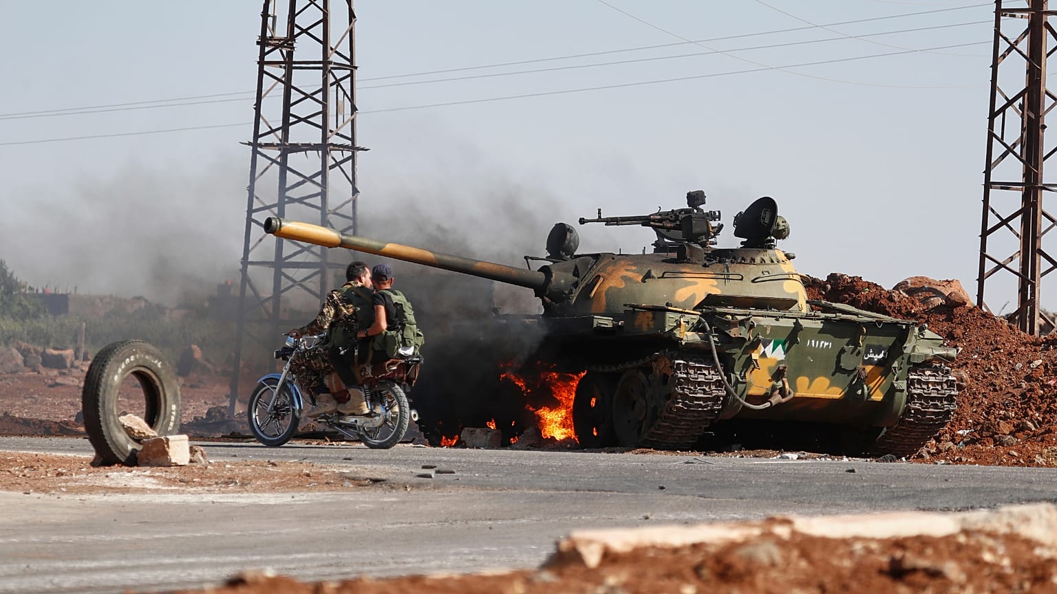Syrian government soldiers on a motorcycle pass by a burning tank on the outskirts of Sweida city, 14 July, 2025