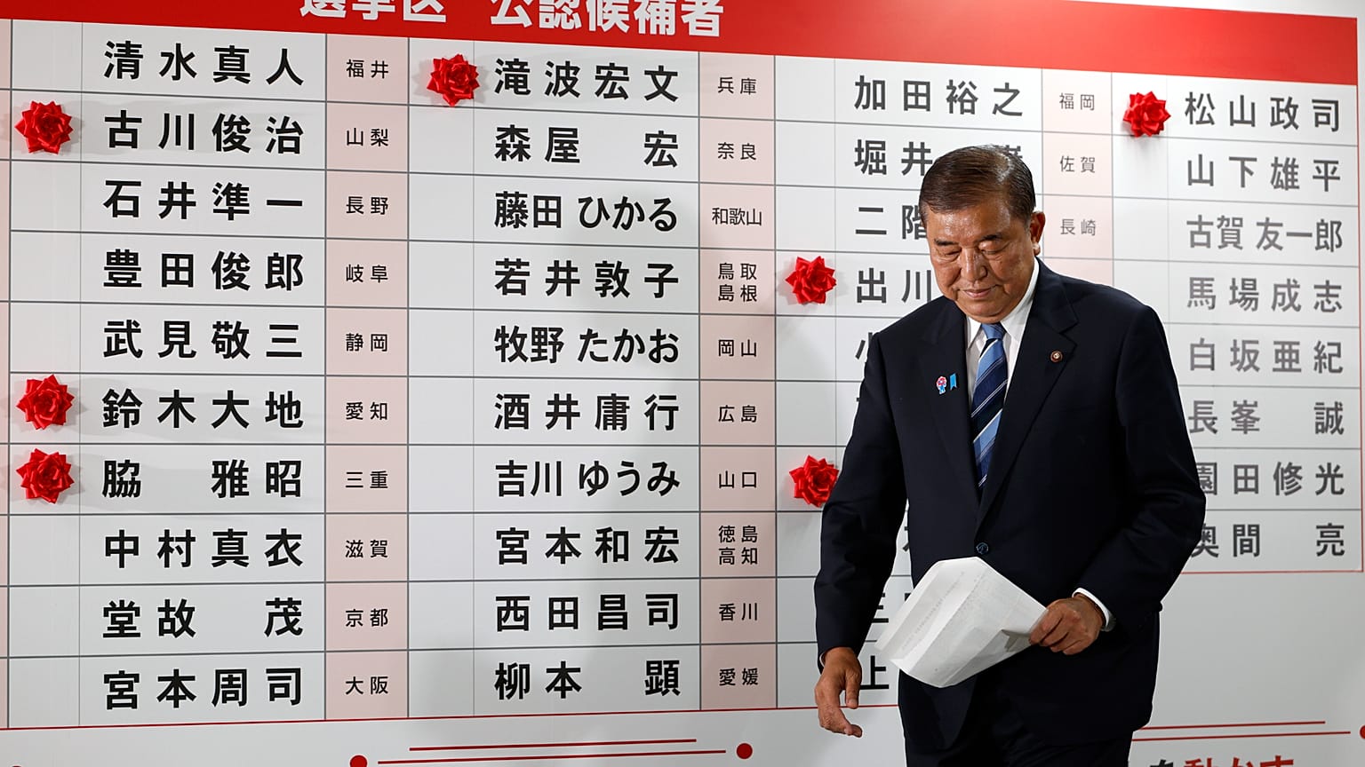 Shigeru Ishiba caminha diante de um quadro com algumas rosas vermelhas de papel que mostra o candidato eleito na sede do LDP em Tóquio, no domingo, 20 de julho de 2025. 
