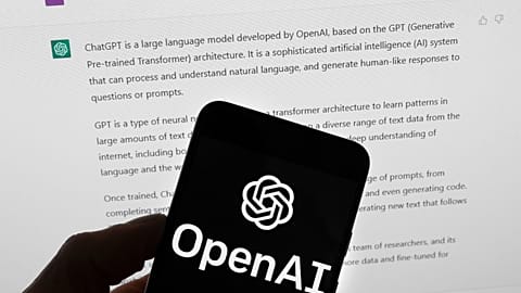 The OpenAI logo is seen on a mobile phone in front of a computer screen which displays output from ChatGPT, March 21, 2023, in Boston
