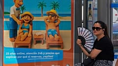 Eine Frau fächelt sich bei heißem Wetter an einer Bushaltestelle neben dem Plakat eines Reisebüros, das Menschen am Strand zeigt, in Madrid, Spanien, am Freitag, 8. August 2025, Luft zu.