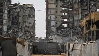 Группа палестинцев сидит в тени своей палатки на фоне разрушенных зданий в жаркий летний день в городе Газа, 12 августа 2025 года.