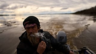 Jake Pogrebinsky fuma un cigarrillo mientras navega con su barca en el río Yukón en busca de troncos para recolectar, el martes 20 de mayo de 2025, en Galena, Alaska.