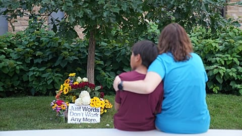 People sit across a memorial near boarded windows damaged in Wednesday's school shooting at Annunciation Catholic Church, Thursday, Aug. 28, 2025, in Minneapolis. 