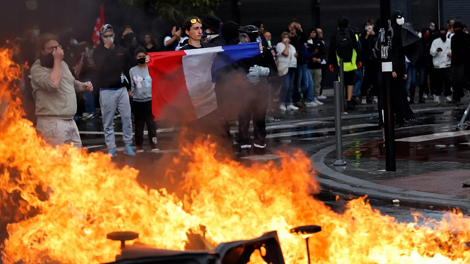 A protester of the "Block Everything" movement displays a French national flag next to burning rubbish bins in Lille, 10 September, 2025