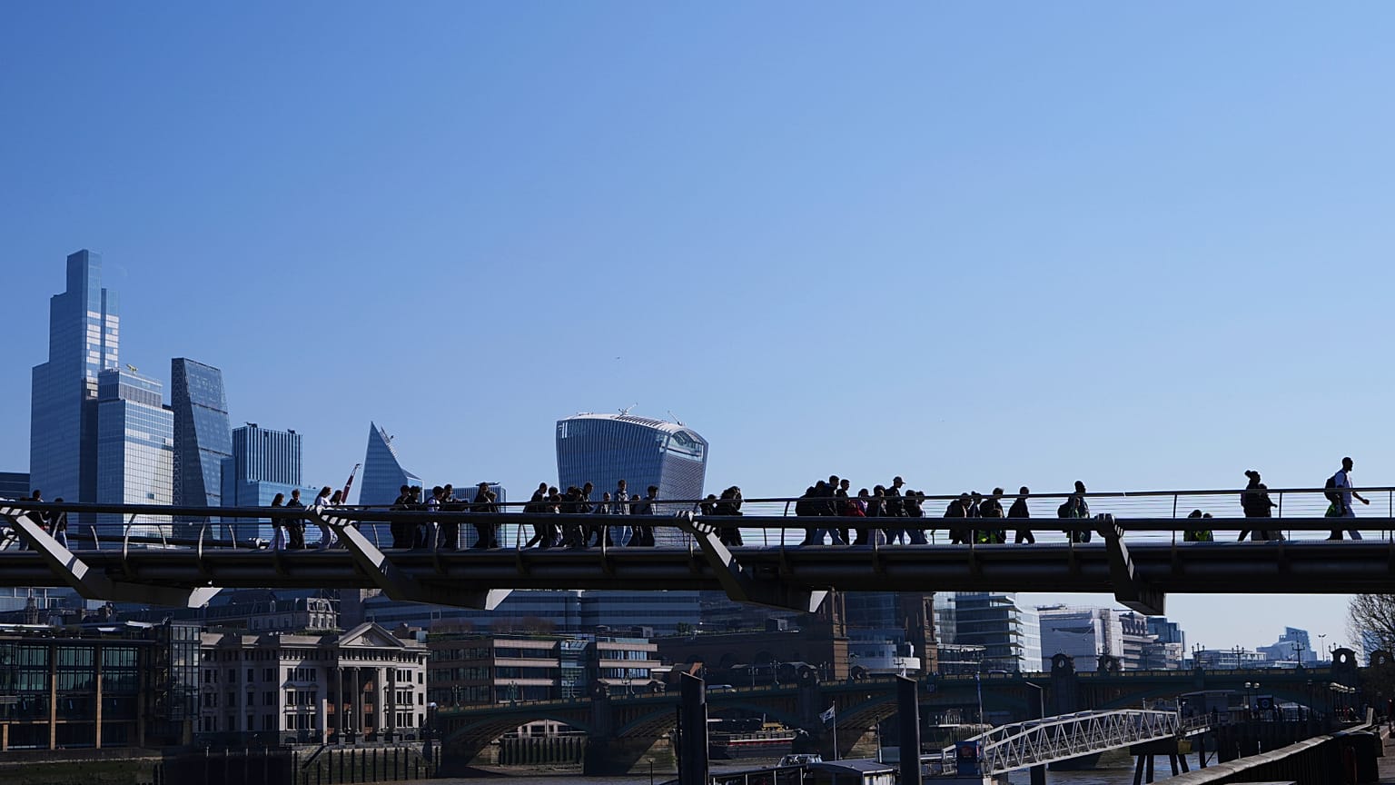 People walk over Millenium Bridge with The City of London buildings in the background. 3 April 2025.