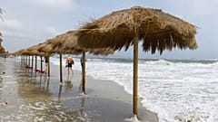 Menschen spazieren an einem menschenleeren Strand entlang, während die Wellen die strohgedeckten Sonnenschirme in Ibiza, Spanien, erreichen, Sonntag, 3. September 2023.