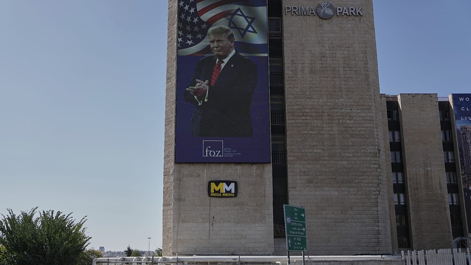 People walk past large outdoor screen displaying an image depicting the U.S. President Donald Trump, in Jerusalem, Saturday, Oct. 11, 2025, after Israel and Hamas agreed to a 