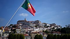 Eine portugiesische Flagge wird auf einer Brücke über den Fluss Mondego gehisst, mit dem Hauptgebäude der Universität von Coimbra im Hintergrund, oben auf dem Hügel, in Coimbra, Portugal
