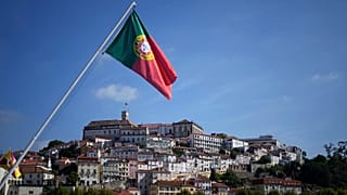 Uma bandeira portuguesa está hasteada numa ponte sobre o rio Mondego, com o edifício principal da Universidade de Coimbra ao fundo, no topo da colina, em Coimbra, Portugal