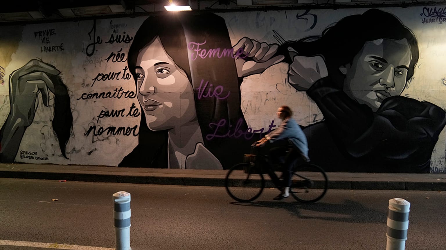 امرأة تركب دراجة أمام لوحة جدارية تحمل توقيع Clacks-one وHeartcraft_Street art، تُصوّر نساء يقدمن على قص شعرهن دعمًا للمتظاهرات الإيرانيات اللواتي يرفضن قيادتهن بعد وفاة شابة 