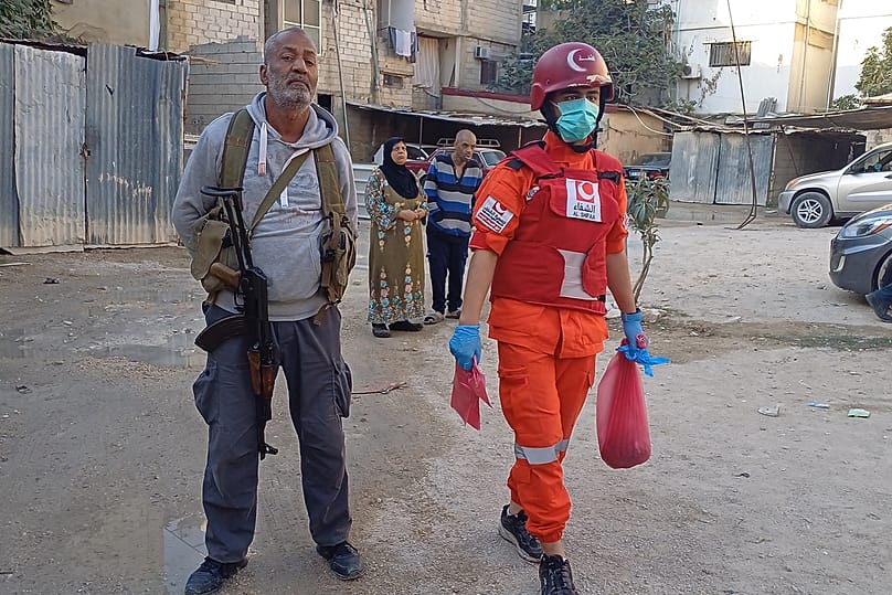  مسلح من حماس، يقف بجوار رجل إنقاذ فلسطيني يحمل حقيبة تحتوي على بقايا جثة، بالقرب من مكان قصف إسرائيلي على مخيم عين الحلوة، الأربعاء 19 نوفمبر/تشرين الثاني 2025. 