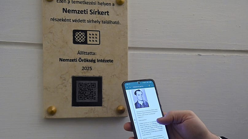 A Nemzeti Sírkertbe sorolt sírhelyeket jelölő okostábla és okostelefonon Buzánszky Jenő fényképe a Szent István-bazilika altemplomában 2025. november 25-én