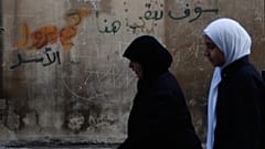 نساء يمشين أمام جدار مع كتابات مكتوبة باللغة العربية "سنبقى هنا ، الأسد"في مدينة حمص ، سوريا ، الجمعة ، نوفمبر. 21, 2025.