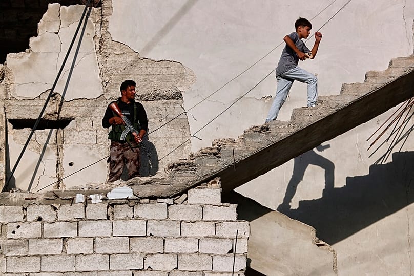 A boy climbs the stairs of a war-damaged building as a security officer stands guard in Zamalka on the outskirts of Damascus, 21 August 2025