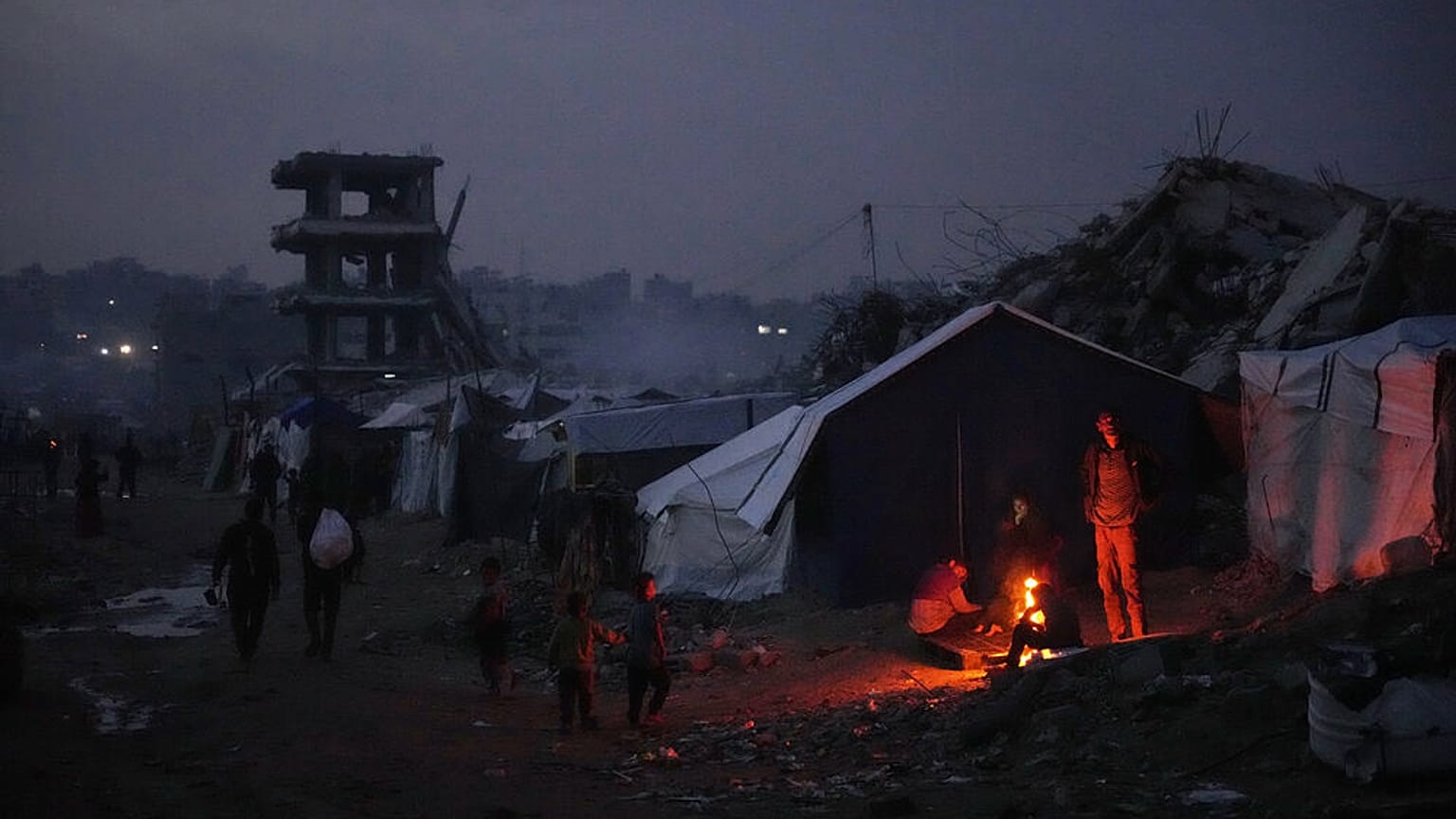 يقف فلسطينيون نازحون قرب نار أوقدوها بجانب خيمتهم وسط الدمار الذي خلّفته العملية الجوية والبرية في مدينة غزة، الجمعة 28 نوفمبر/تشرين الثاني 2025. (أ ب / عبد الكريم هنا)