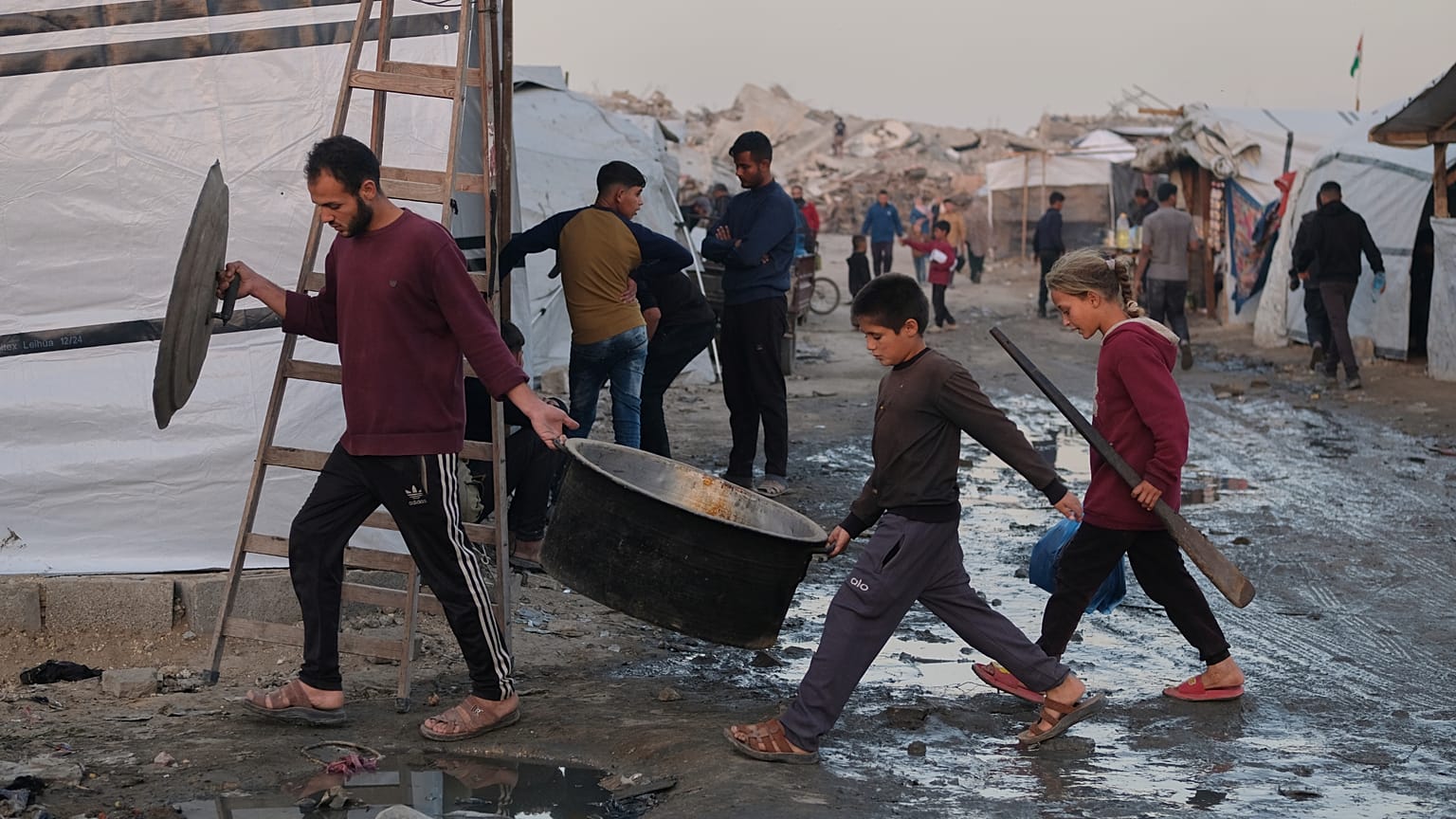 Palestinianos deslocados caminham por trilhas lamacentas enquanto um homem e crianças carregam utensílios de cozinha num campo em Jabalia, no norte da Faixa de Gaza
