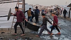 Palestinianos deslocados caminham por trilhas lamacentas enquanto um homem e crianças carregam utensílios de cozinha num campo em Jabalia, no norte da Faixa de Gaza