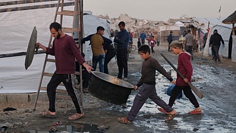 Palestinianos deslocados caminham por trilhas lamacentas enquanto um homem e crianças carregam utensílios de cozinha num campo em Jabalia, no norte da Faixa de Gaza