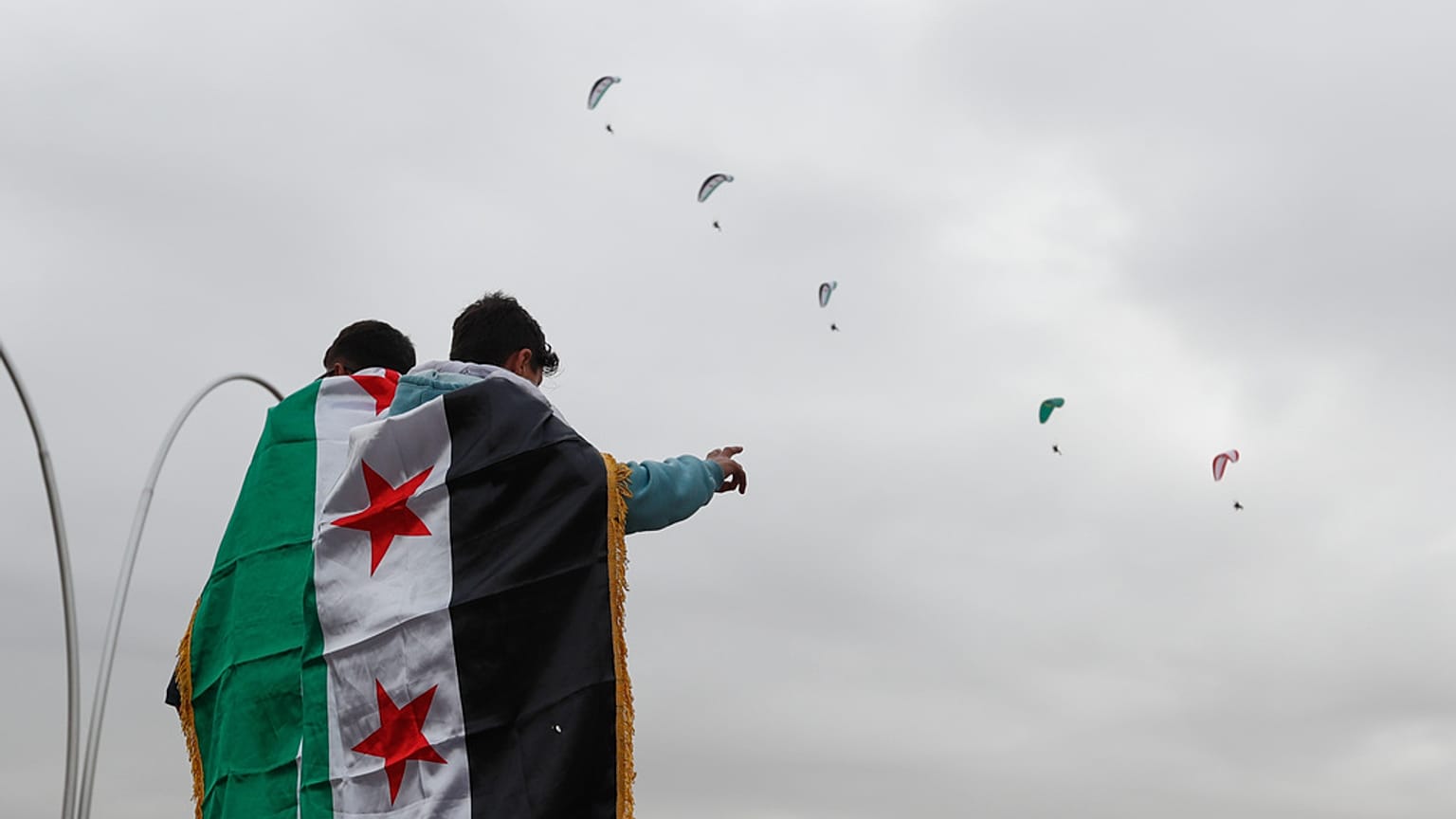 أشخاص ملفوفون بالأعلام السورية يشاهدون طائرات شراعية تحلق فوق رؤوسهم، دمشق، سوريا، الاثنين 8 ديسمبر/كانون الأول 2025.