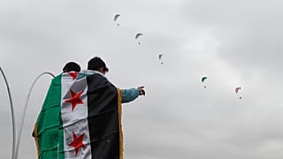 أشخاص ملفوفون بالأعلام السورية يشاهدون طائرات شراعية تحلق فوق رؤوسهم، دمشق، سوريا، الاثنين 8 ديسمبر/كانون الأول 2025.