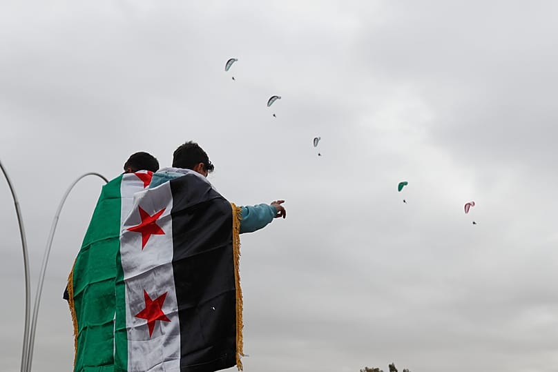 People wrapped in Syrian flags watch paragliders fly overhead during the first anniversary of the ousting of former President Bashar al-Assad, in Damascus, 8 December 2025