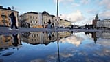 I passanti nella piazza si riflettono in una pozzanghera a Helsinki, in Finlandia, il 14 novembre 2025.