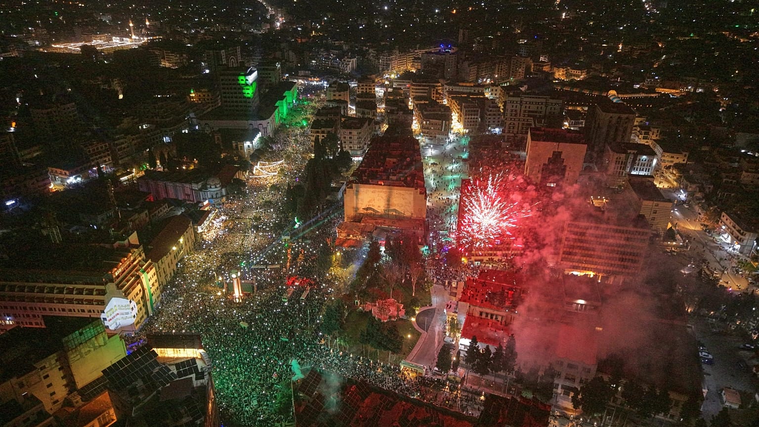 Fogo de artifício e luzes iluminam o céu, pessoas enchem a Praça do Relógio em Homs, Síria ocidental, seg., 8 dez. 2025, marcando o 1.º aniversário do derrube de