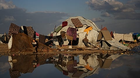 A Palestinian boy stands next to his mother washing clothes as mattresses hang to dry over their tent in a makeshift camp