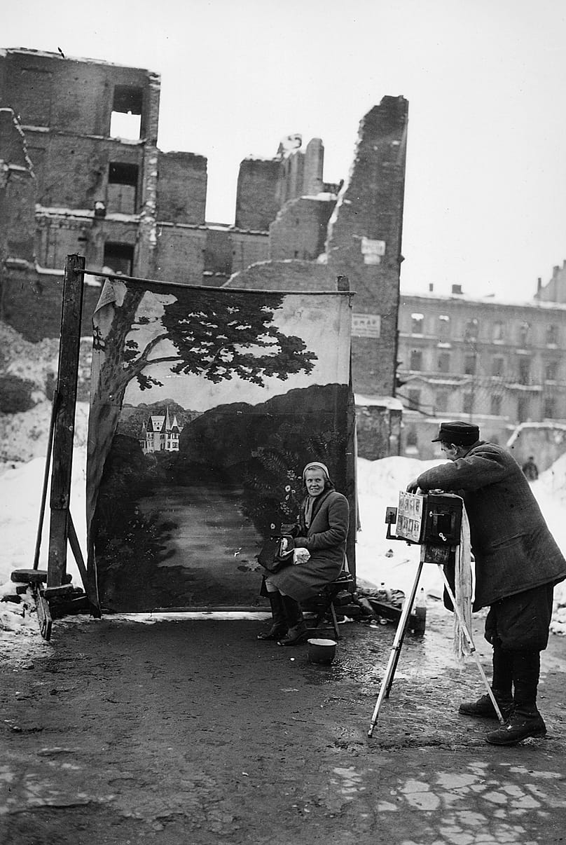 Um fotógrafo de rua usa o seu próprio fundo (backdrop) para obscurecer as ruínas de Varsóvia destruídas durante a Segunda Guerra Mundial, enquanto tira o retrato de uma mulher; novembro de 1946