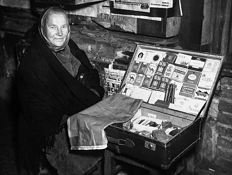 Uma mulher idosa senta-se num banco, vendendo todas as marcas mais populares de cigarros ingleses e americanos numa rua de Varsóvia, a 17 de dezembro de 1946. 