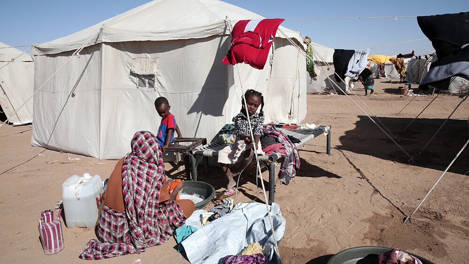 امرأة سودانية نازحة من الفاشر تغسل الملابس خارج خيمة عائلتها بينما يجلس الأطفال بالقرب منها في مخيم الأفضح الذي تم إنشاؤه حديثًا في الدبة، بولاية الشمالية في السودان