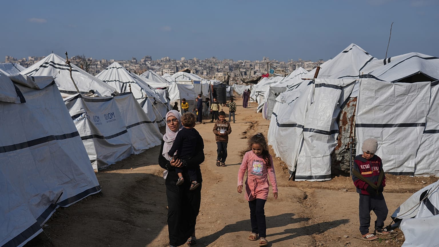 امرأة فلسطينية تحمل طفلاً أثناء سيرها بين خيام أقيمت للنازحين في مدينة غزة، يوم الأربعاء 17 ديسمبر 2025. (تصوير: جيهاد الشرافي/أسوشيتد برس)