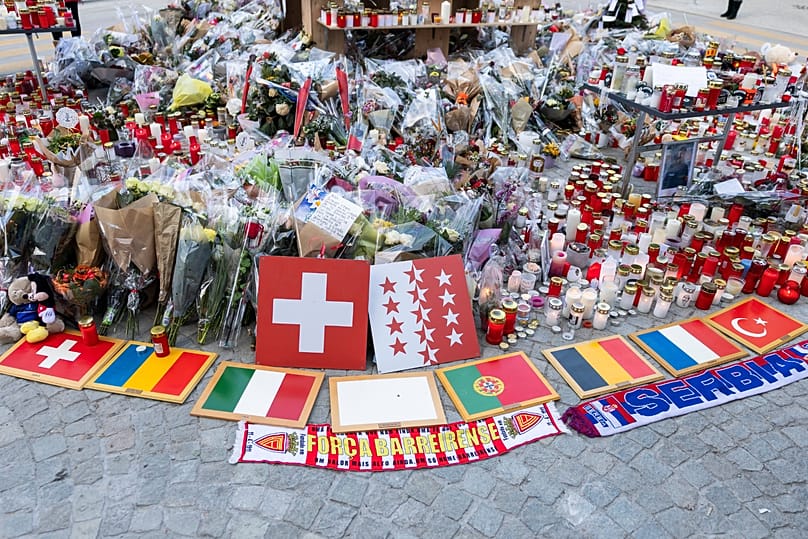 People stand around floral tributes and candles placed outside the sealed off Le Constellation bar in Crans-Montana, 6 January, 2026