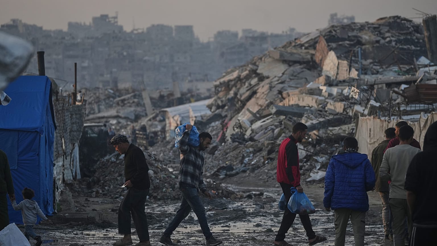 فلسطينيون يمشون أمام مخيم خيام وسط مبان دمرتها العمليات الجوية والبرية الإسرائيلية في مدينة غزة الثلاثاء, يناير. 6, 2026