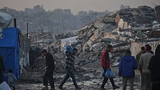 فلسطينيون يمشون أمام مخيم خيام وسط مبان دمرتها العمليات الجوية والبرية الإسرائيلية في مدينة غزة الثلاثاء, يناير. 6, 2026