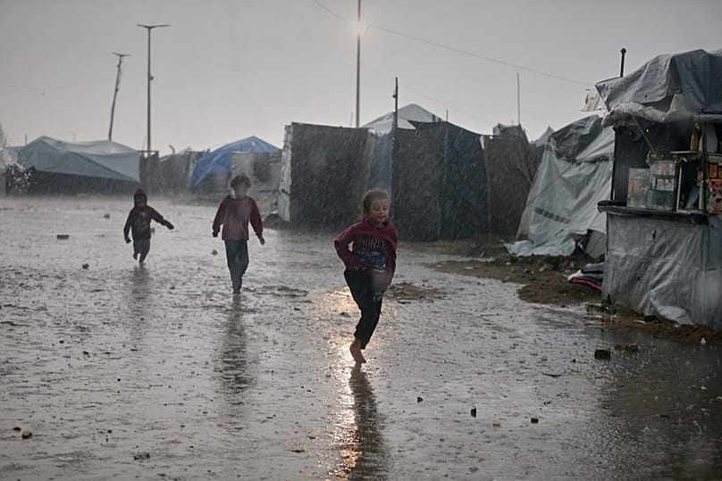  أطفال يركضون تحت المطر أمام مخيم خيام في مدينة غزة، يوم الاثنين 15 ديسمبر/كانون الأول 2025.