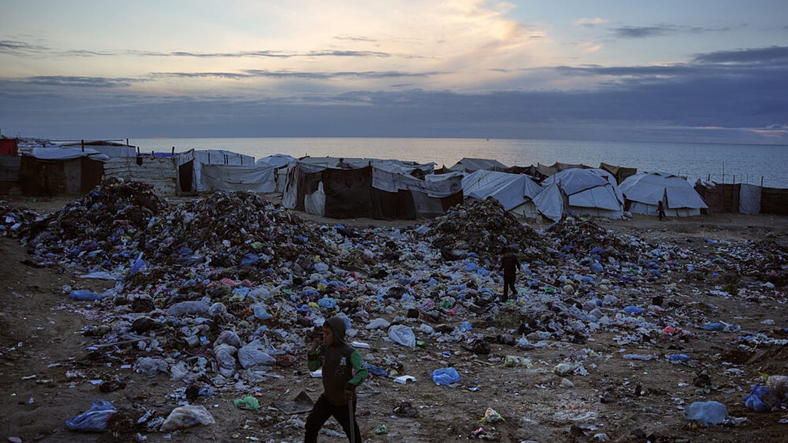 أطفال يسيرون فوق أكوام من النفايات في مخيم خيام مؤقت للنازحين الفلسطينيين على أحد شواطئ مدينة دير البلح وسط قطاع غزة، الجمعة 16 جانفي/يناير 2026.