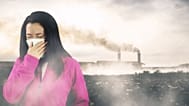 امرأة تضع كمامة فيما يتصاعد الدخان من المصانع في الخلفية