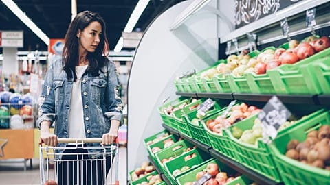 In this photo, a young woman walks along the fruit and vegetable row in a supermarket, pushing a shopping trolley.