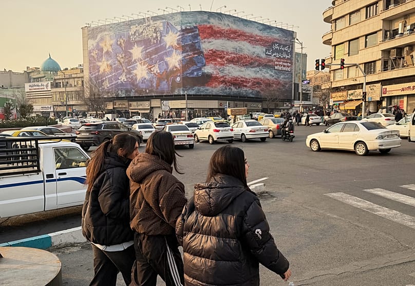 People walk in front a billboard with graphic showing a US aircraft carrier with damaged fighter jets on its deck in Tehran, 25 January, 2026