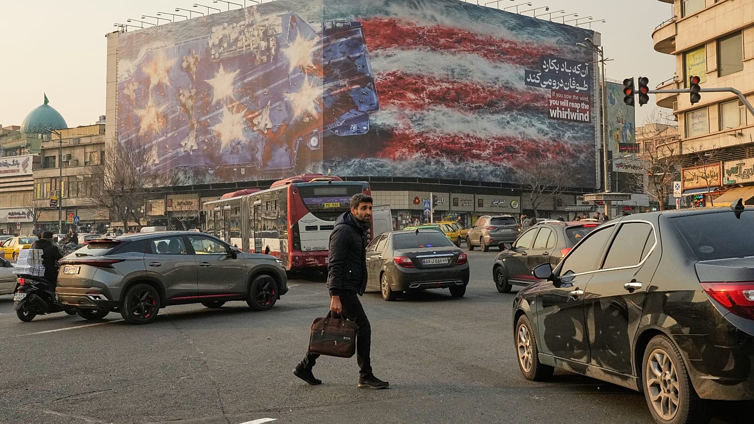 A man walks among vehicles in front of a billboard depicting a damaged U.S. aircraft carrier with disabled fighter jets on its deck.