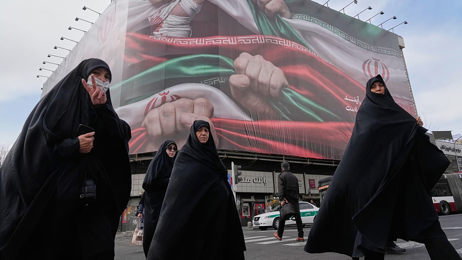 نساء، إحداهن تلوح بيد النصر، يعبرن الشارع تحت لافتة ضخمة تظهر أيديهن تحمل الأعلام الوطنية الإيرانية بقوة كدليل على الوطنية، في طهران، إيران، 14 يناير 2026.