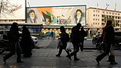 أشخاص يسيرون أمام جدارية تُصوّر مؤسس الثورة الإيرانية الراحل آية الله الخميني (يمينًا)، إلى جانب عناصر من قوات الباسيج شبه العسكرية والمرشد الأعلى آية الله علي خامنئي