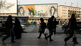 أشخاص يسيرون أمام جدارية تُصوّر مؤسس الثورة الإيرانية الراحل آية الله الخميني (يمينًا)، إلى جانب عناصر من قوات الباسيج شبه العسكرية والمرشد الأعلى آية الله علي خامنئي