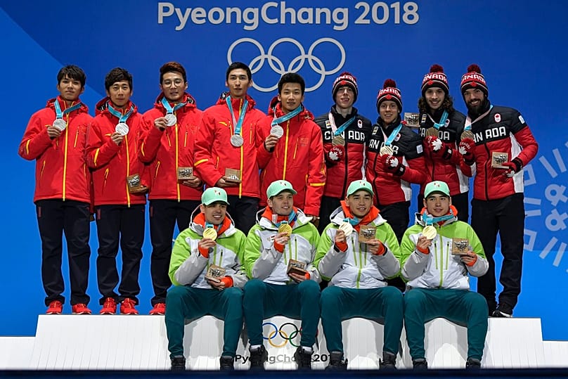 Burján Csaba, Knoch Viktor, Liu Shaolin Sándor és Liu Shaoang (ülve) az olimpiai dobogón, mögöttük a második helyezett kínai és a harmadik kanadai csapat 2018. február 23-án. 