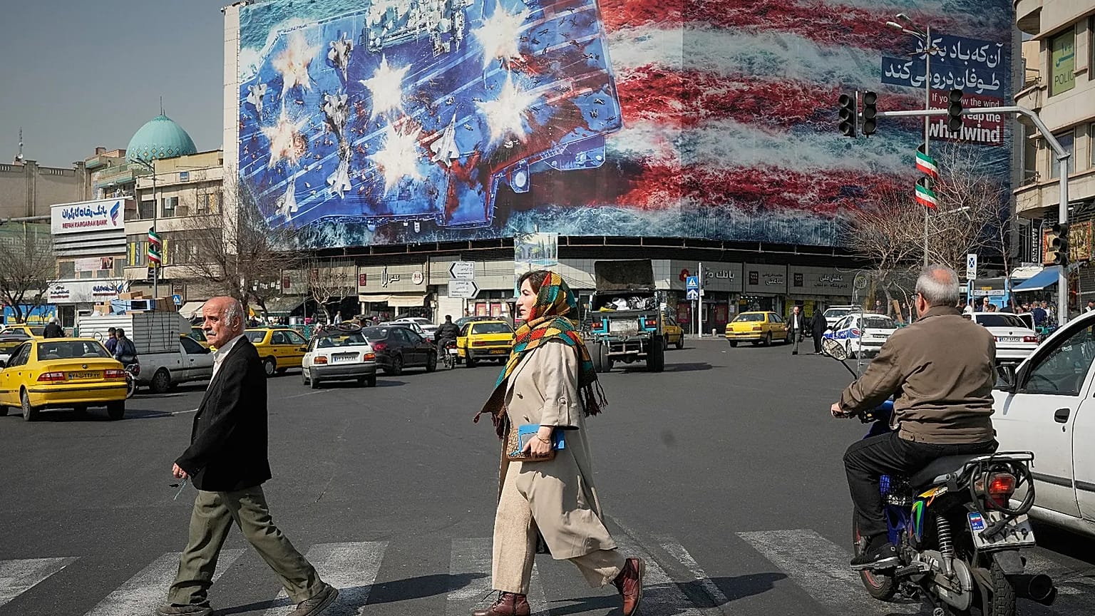 Pedestrians walk past a billboard depicting a U.S. aircraft carrier with damaged fighter jets on its deck in Tehran, Iran, Sunday, Feb. 22, 2026.