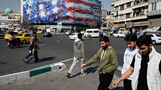 يمر المشاة أمام لوحة إعلانية تصور حاملة طائرات أمريكية وعلى سطحها طائرات مقاتلة تالفة ولافتة باللغتين الفارسية والإنجليزية كتب عليها "إذا زرعت الريح، فسوف تحصد الزوبعة"، طهران