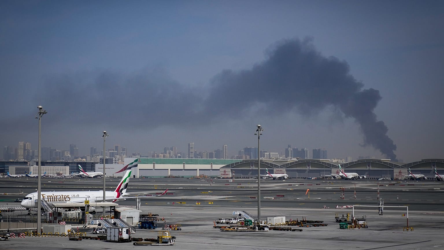 A plume of smoke caused by an Iranian strike is seen in the background as Emirates planes are parked at Dubai International Airport after its closure
